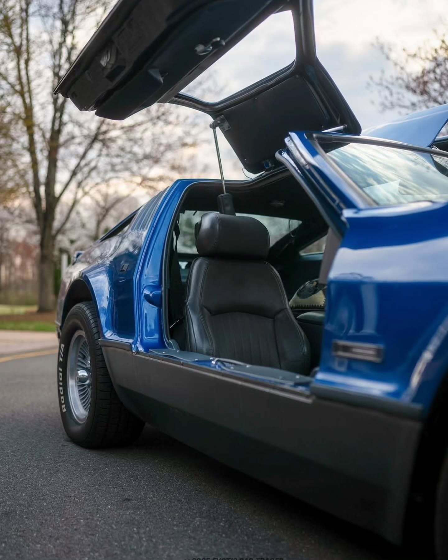 نمای جانبی خودروی کلاسیک بریکلین SV-1 با درهای باز شده رو به بالا و بدنه آکریلیک نارنجی رنگ که طراحی ایمن و آیرودینامیک آن را نشان می‌دهد.